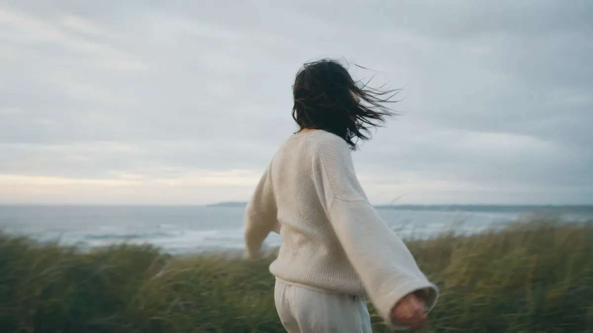 A woman in a cream sweater turns on a windswept coastal dune, her dark hair blowing across her face, the grey sea behind.